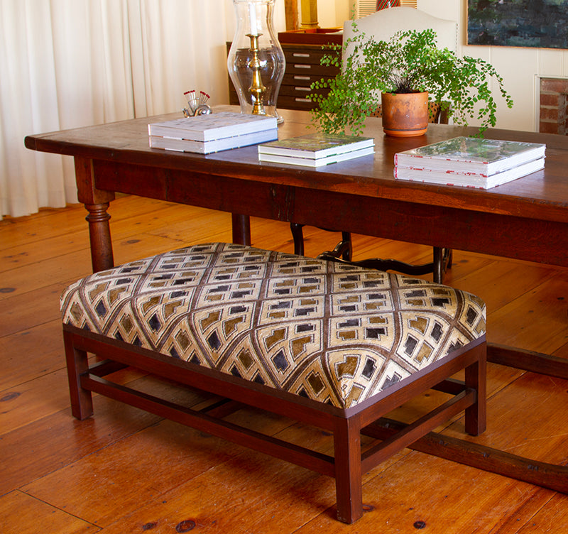 Ottoman upholstered in sand and cognac Kuba cloth epingle velvet with espresso trim, measuring 42”W x 24”D x 17”H in front of a wood table.